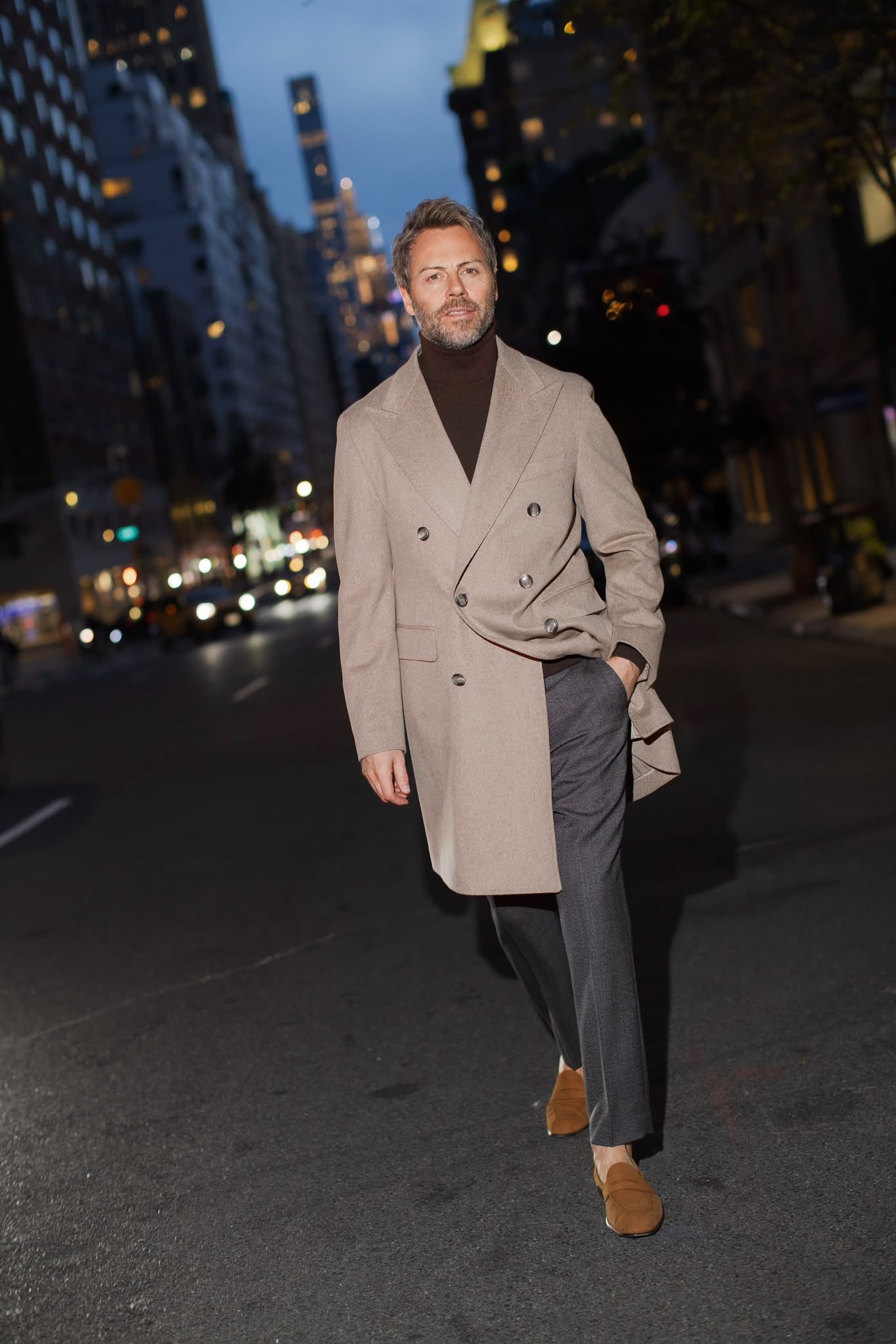 Gilberto Ramerini in a camel overcoat and brown turtleneck, on a city street at dusk